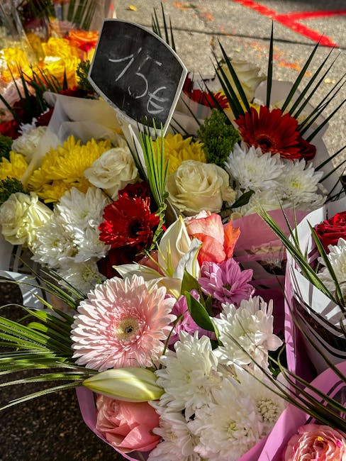 A vibrant bouquet of fresh flowers displayed outdoors, featuring large pink gerbera daisies, white roses, red gerbera daisies, and white chrysanthemums, all arranged with long green palm leaves and ot