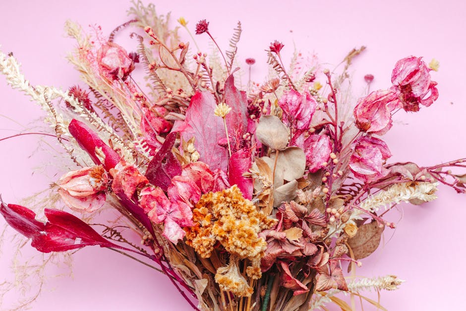 A bouquet of dried flowers arranged on a soft pink background, featuring pink and red roses with wilted petals, pale beige and cream-colored grasses, and small yellow and red accents. The dried bouque