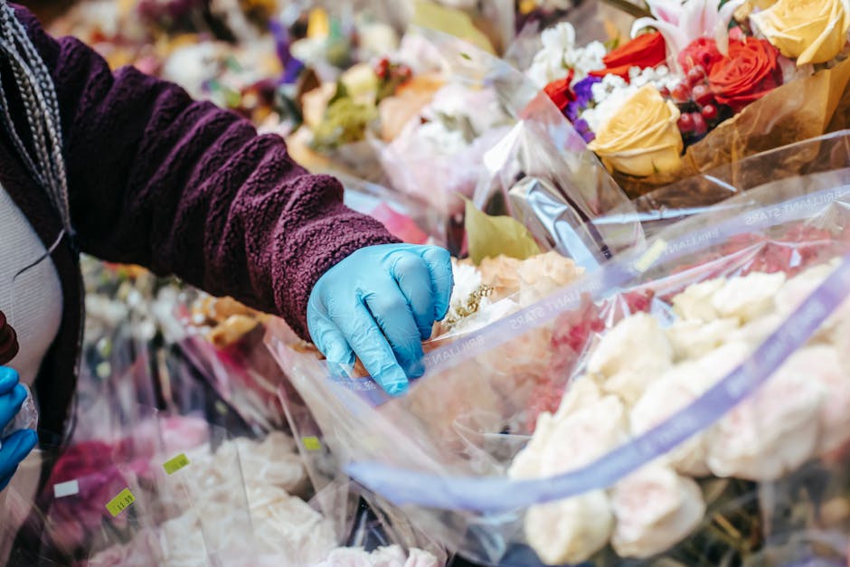 A person wearing a purple velvet sweater and blue gloves is selecting flowers from large, clear plastic bags filled with fresh, colorful blooms at a florist's shop. The arrangement includes roses in s