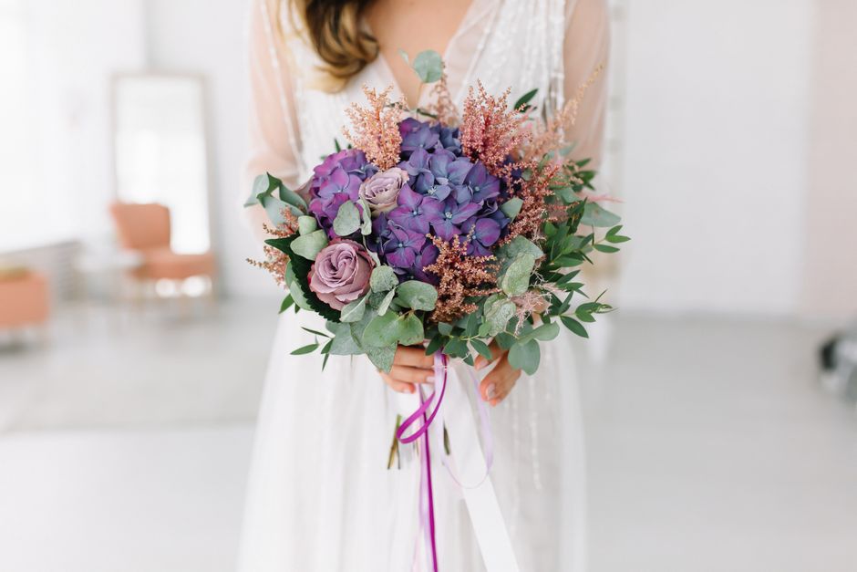 A bridal bouquet featuring predominantly purple and lavender roses, accented with large purple hydrangeas, light pink astilbe, and green eucalyptus leaves, all arranged in a rounded, lush style. The b