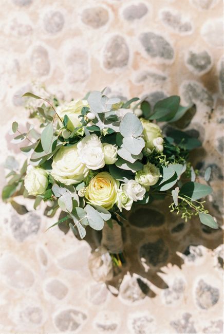 A bouquet of fresh flowers featuring predominantly white ranunculus and roses, accented with light green eucalyptus leaves and jasmine blooms, arranged in a compact, rounded style. The bouquet is wrap