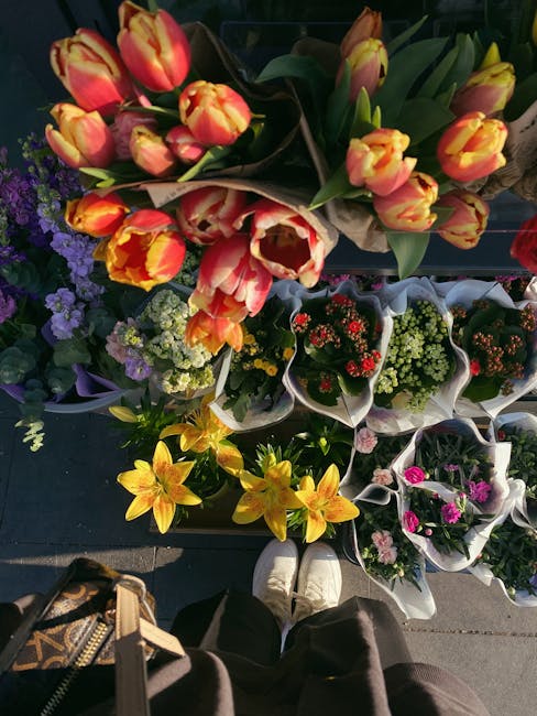 A vibrant display of fresh flowers arranged in bouquets and potted plants, including orange and yellow tulips with green stems and leaves, lavender purple statice, yellow lilies, and a variety of othe