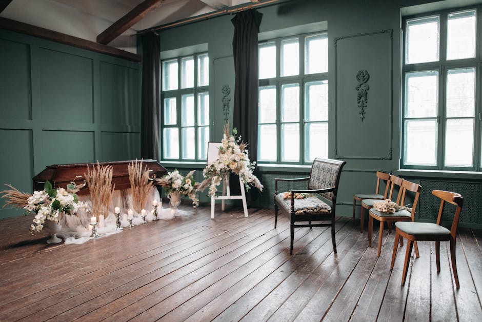 A floral arrangement featuring white roses, peonies, and lilies, accented with tall, fluffy pampas grass and smaller white blossoms, is displayed on a white easel in a softly lit room with dark green-