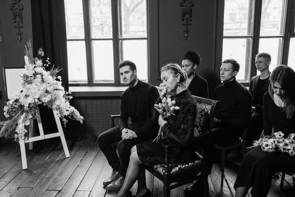 A group of six people, including women and men, seated indoors during a funeral ceremony, with five of them holding floral arrangements or bouquets. The woman in the center, with short hair, is holdin