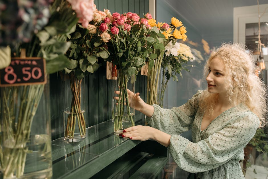 A woman with curly blonde hair and fair skin selects a bouquet of mixed fresh flowers in a floral shop. She is wearing a light, floral-patterned blouse with long sleeves and has red nail polish. She i